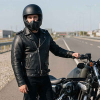 Man in black Leather Moto Jacket and helmet standing next to a motorcycle on an open road.